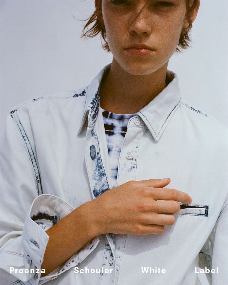 Close-up shot of a woman with short brown hair wearing a white, button-down denim shirt, posing indoors against a white backdrop.