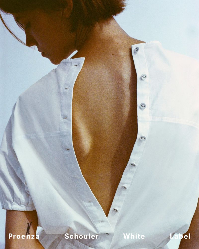 Back shot of a woman in an unbuttoned white shirt, revealing her bare back and shoulders against a neutral studio backdrop.