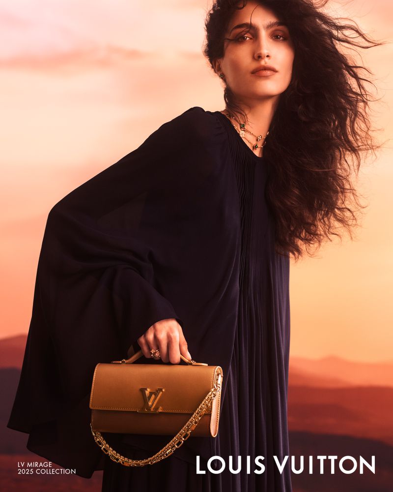 Model with dark wavy hair wearing a black dress holding a tan handbag against a sunset sky