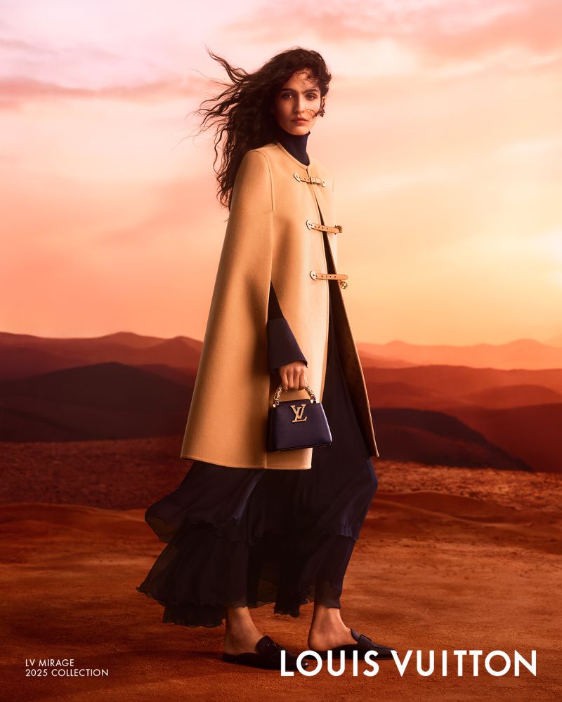 Fashion model showcases Louis Vuitton cape coat and handbag in a desert landscape against an orange sunset sky, fashion editorial.