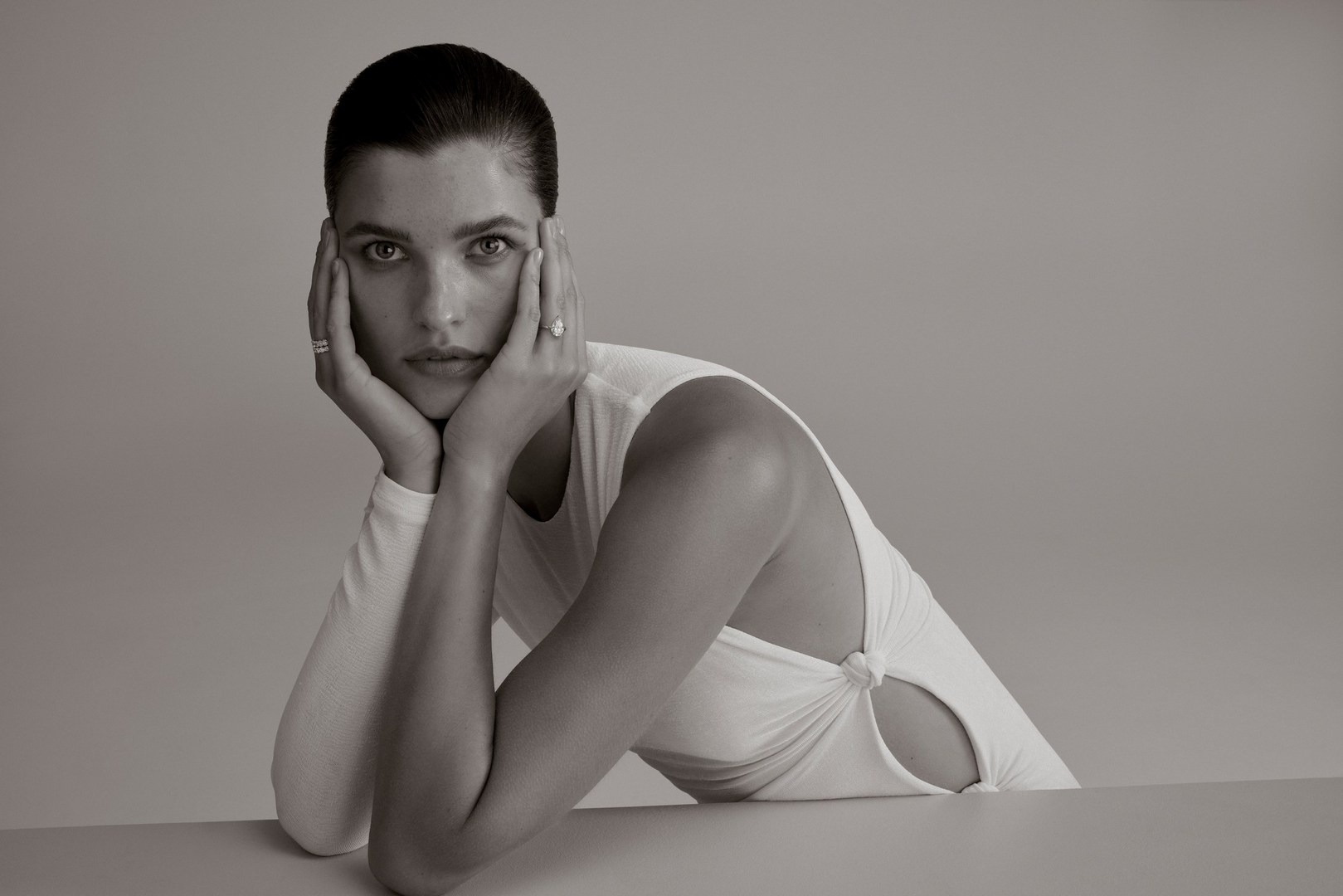 Portrait of an adult female with hands on her face, wearing a white top, against a grey background, in a studio environment.
