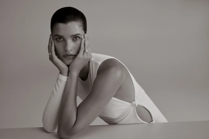 Portrait of an adult female with hands on her face, wearing a white top, against a grey background, in a studio environment.