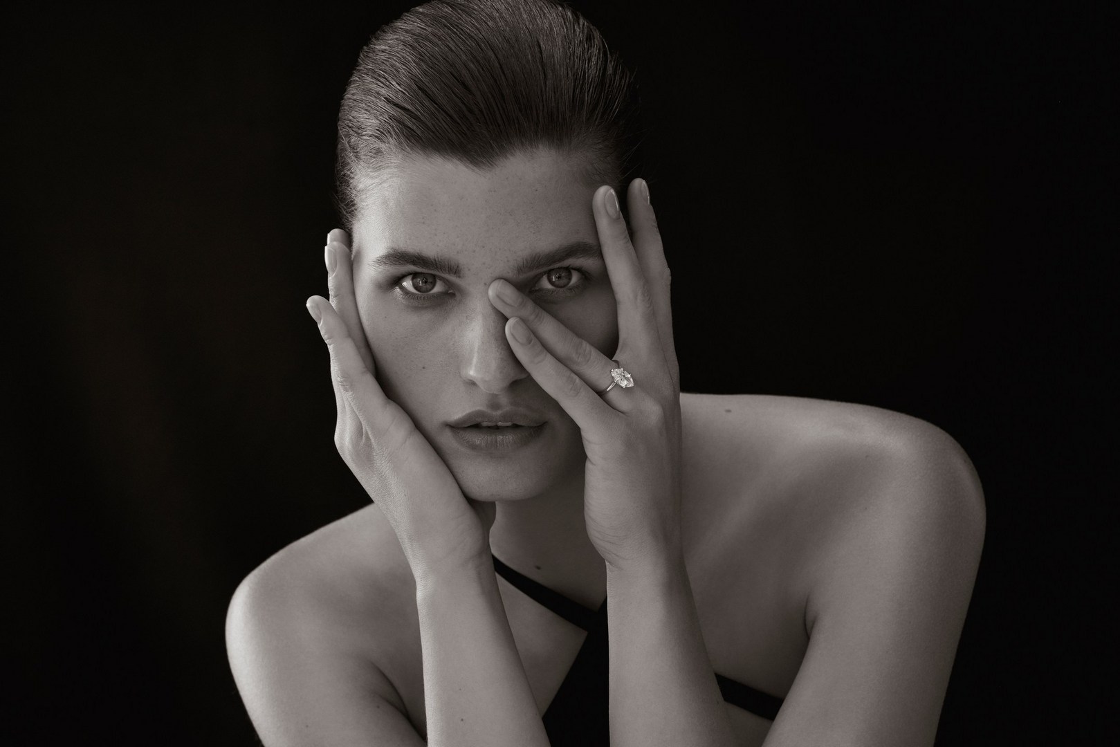 Striking black-and-white portrait of an adult with hands framing their face, showcasing a prominent ring, set against a dark background.