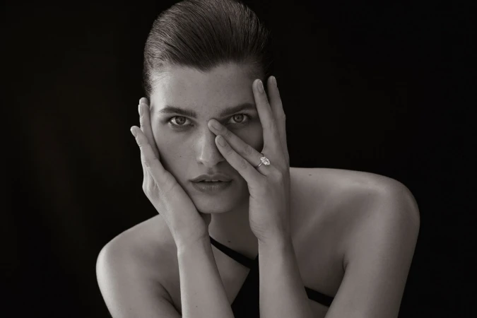 Striking black-and-white portrait of an adult with hands framing their face, showcasing a prominent ring, set against a dark background.