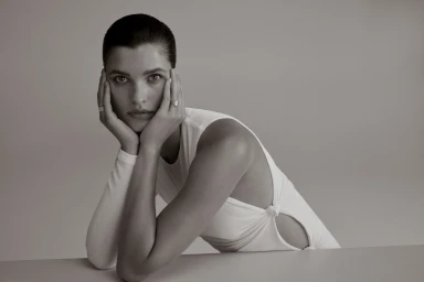 Woman in white sleeveless top leaning forward with hands resting on her face against a plain grey studio backdrop