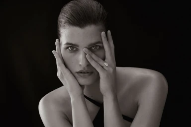 Close-up portrait of a woman with slicked-back hair holding her hands to her face, wearing a diamond ring.