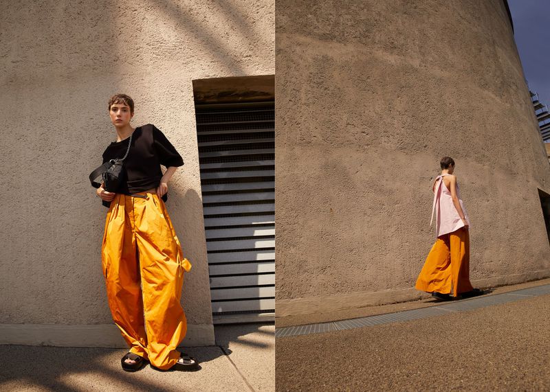 Fashionable young adult with short hair poses in orange pants and black shirt in front of a building on a sunny day.