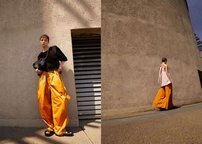 Fashionable young adult with short hair poses in orange pants and black shirt in front of a building on a sunny day.