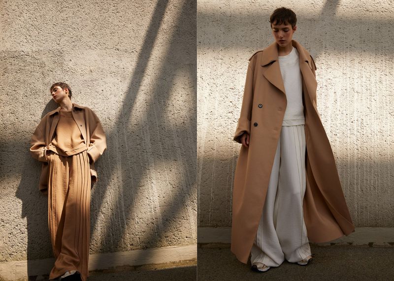 Two women in neutral-toned outfits pose against a textured wall in natural light for a fashion editorial, showcasing contemporary style.