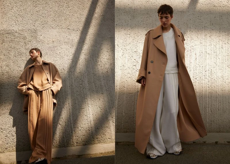 Two women in neutral-toned outfits pose against a textured wall in natural light for a fashion editorial, showcasing contemporary style.