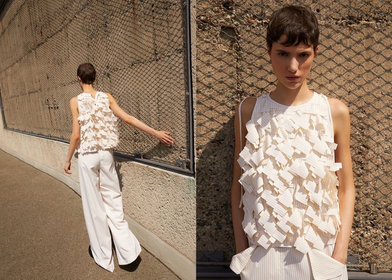 Fashion shot of a woman in an off-white top with layered fabric and wide-leg pants, posing against a textured concrete wall and metal grating.