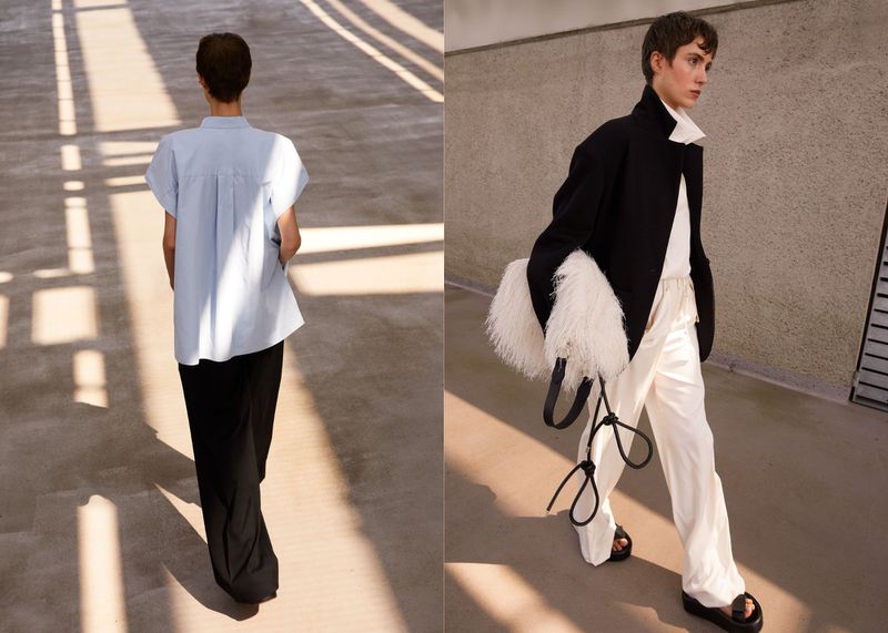Two fashion models showcase modern styles in an urban setting with natural light. One model faces away in black pants and light shirt.