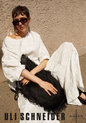 Model wearing dark sunglasses and a white textured dress holding a black fringed bag against a textured wall.