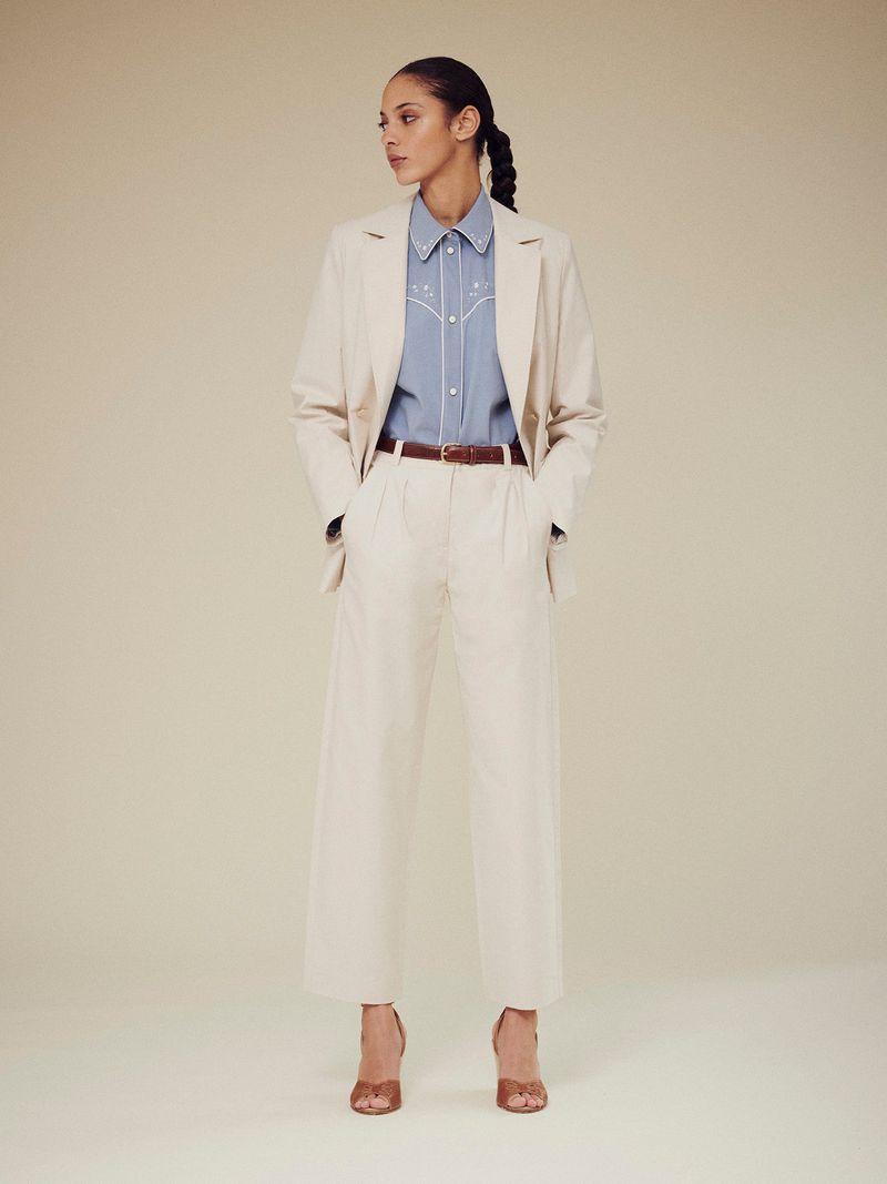 Full-length shot of a woman in a neutral-toned blazer and wide-leg trousers, paired with a blue button-down and brown heels, on a beige background.