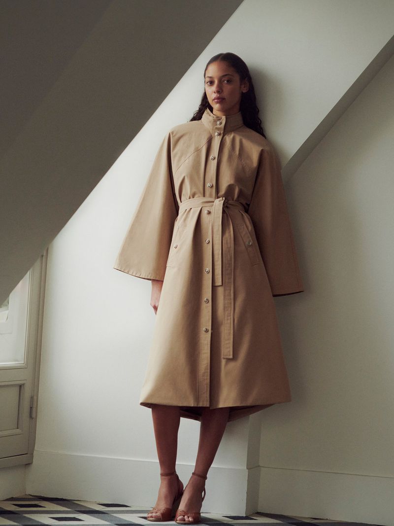 A woman in a long beige trench coat stands against a wall with a patterned floor. Fashion shot with soft, diffused lighting.