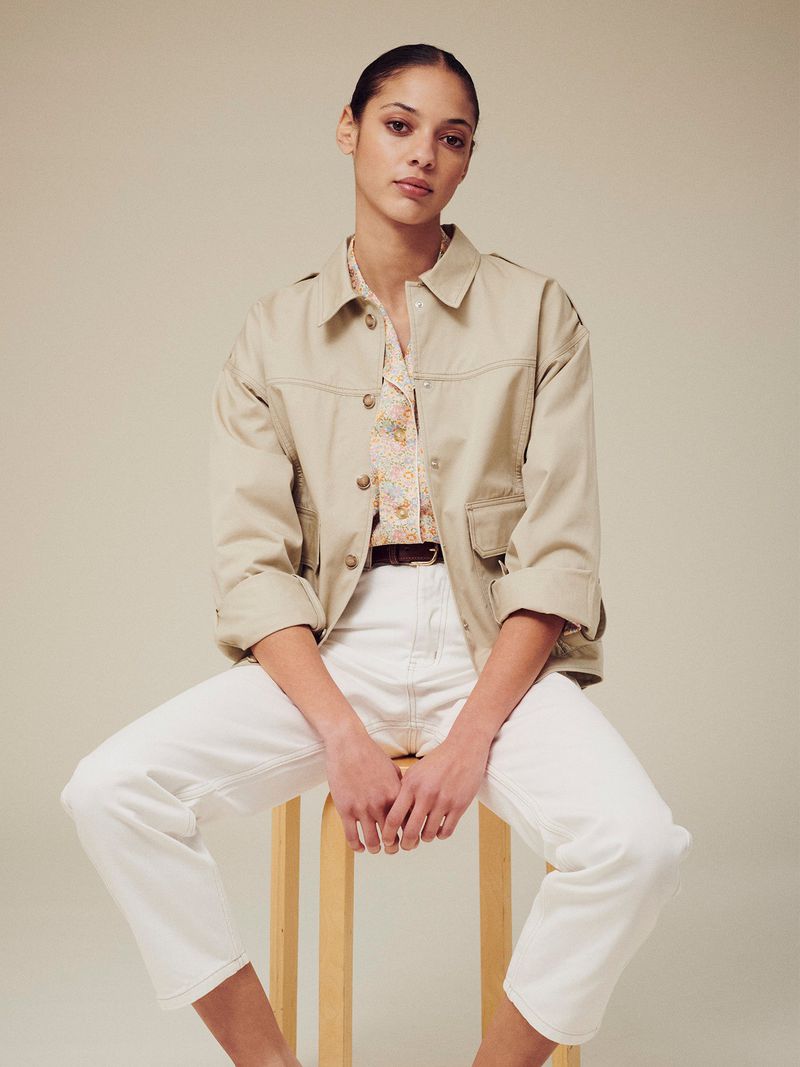 Model seated on a wooden stool wearing a beige utility jacket, patterned shirt, and white trousers against a neutral backdrop.