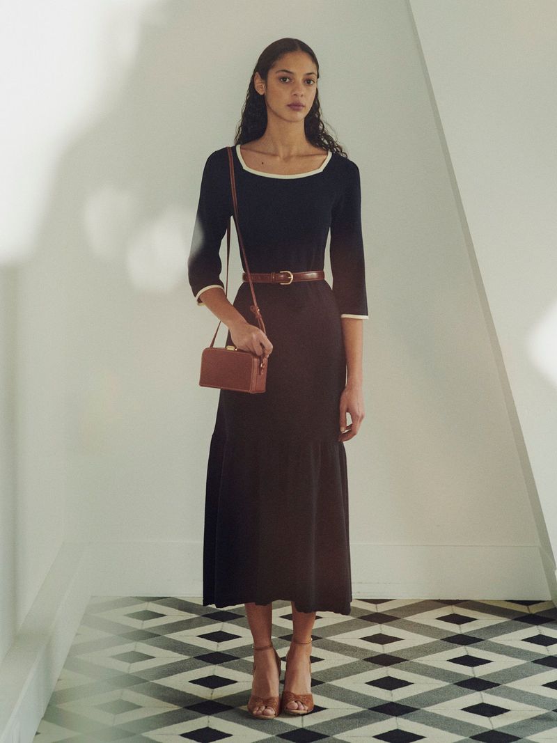 A slender, dark-haired woman poses in a studio wearing a dark blue, calf-length dress with a brown belt and purse with brown heels.