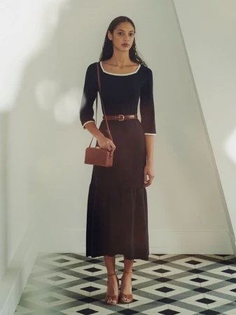 A slender, dark-haired woman poses in a studio wearing a dark blue, calf-length dress with a brown belt and purse with brown heels.