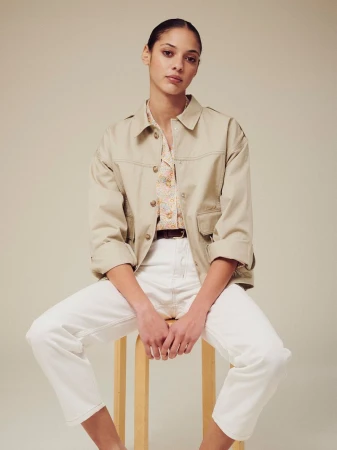 Model seated on a wooden stool wearing a beige utility jacket, patterned shirt, and white trousers against a neutral backdrop.