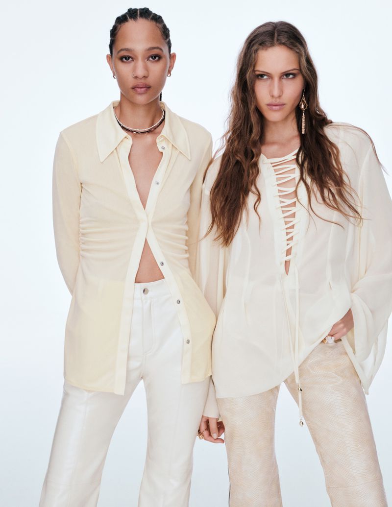 Two women pose in a studio, wearing sheer off-white shirts and pants in a fashion shot with a white backdrop and soft, diffused lighting.