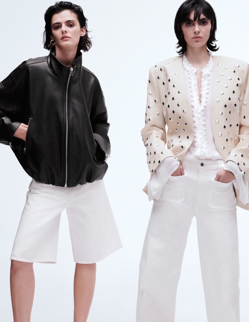 Two adult women pose in a bright photo studio wearing fashionable jackets, white shorts, and wide-leg white pants in a fashion editorial shot.