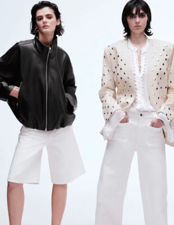 Two adult women pose in a bright photo studio wearing fashionable jackets, white shorts, and wide-leg white pants in a fashion editorial shot.