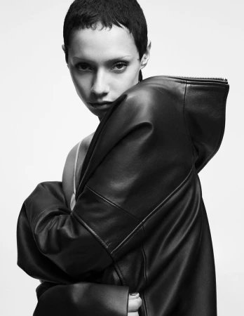 Short-haired person wearing a black leather hooded jacket against a plain white background