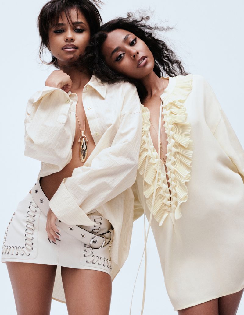 Two dark-skinned women pose in pale clothing against a light backdrop. One wears an open shirt and mini-skirt, the other a ruffled dress.