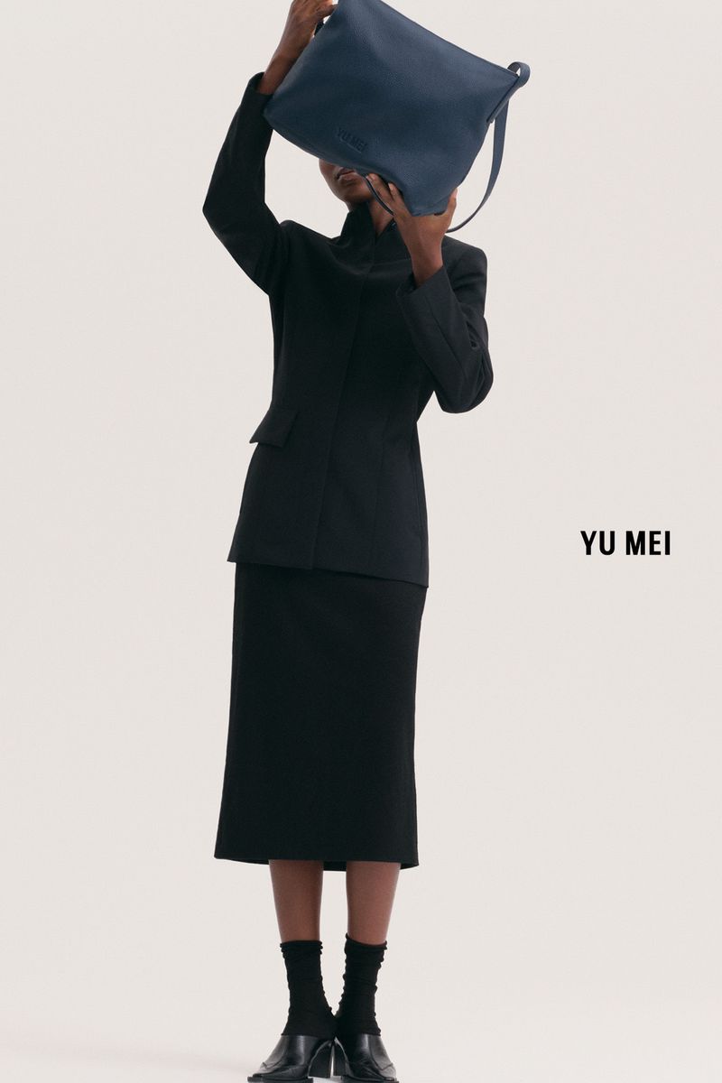 Model holding a blue leather handbag over their face wearing a black suit jacket and matching skirt against white