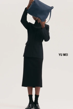 Model holding a blue leather handbag over their face wearing a black suit jacket and matching skirt against white
