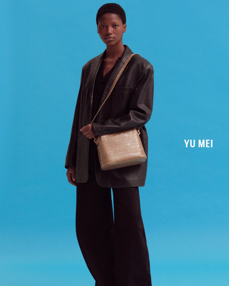 Fashion shot of an African-American woman in a black blazer and pants holding a tan purse against a bright blue studio backdrop.