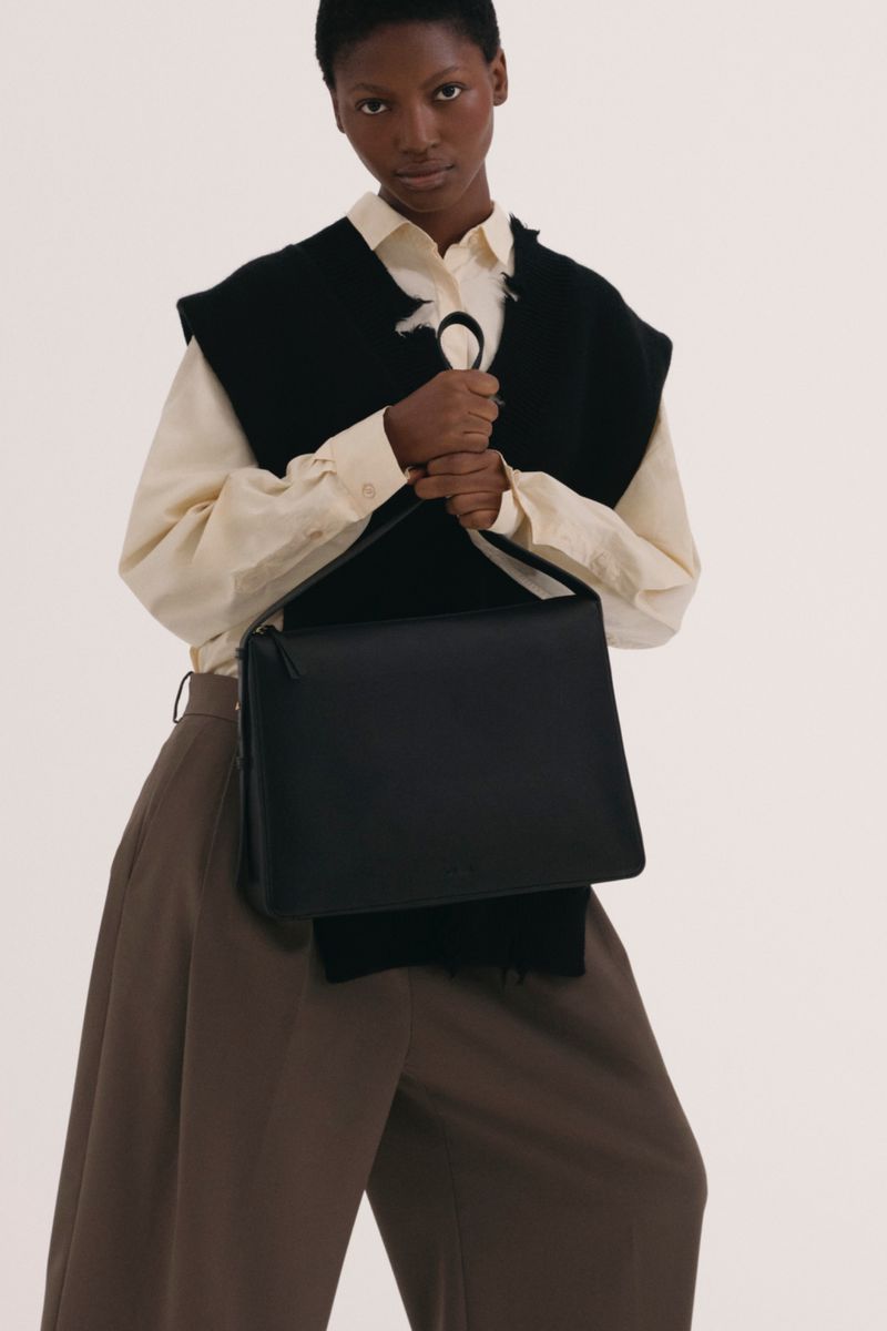 Fashionable adult woman presents a black satchel handbag in a studio shot, accessorized with a sleeveless black vest and loose brown trousers.