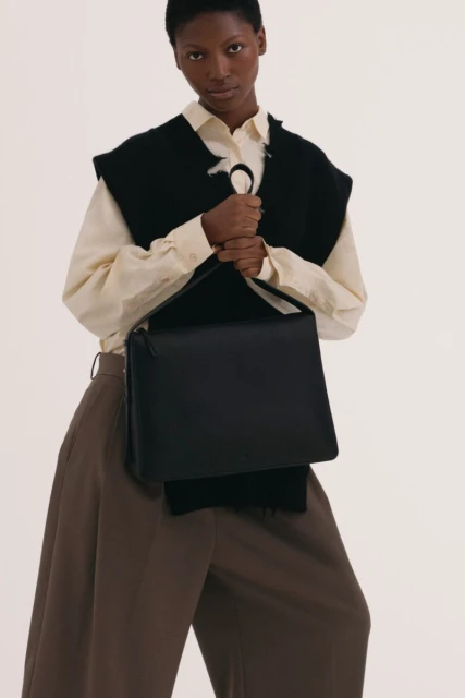 Fashionable adult woman presents a black satchel handbag in a studio shot, accessorized with a sleeveless black vest and loose brown trousers.