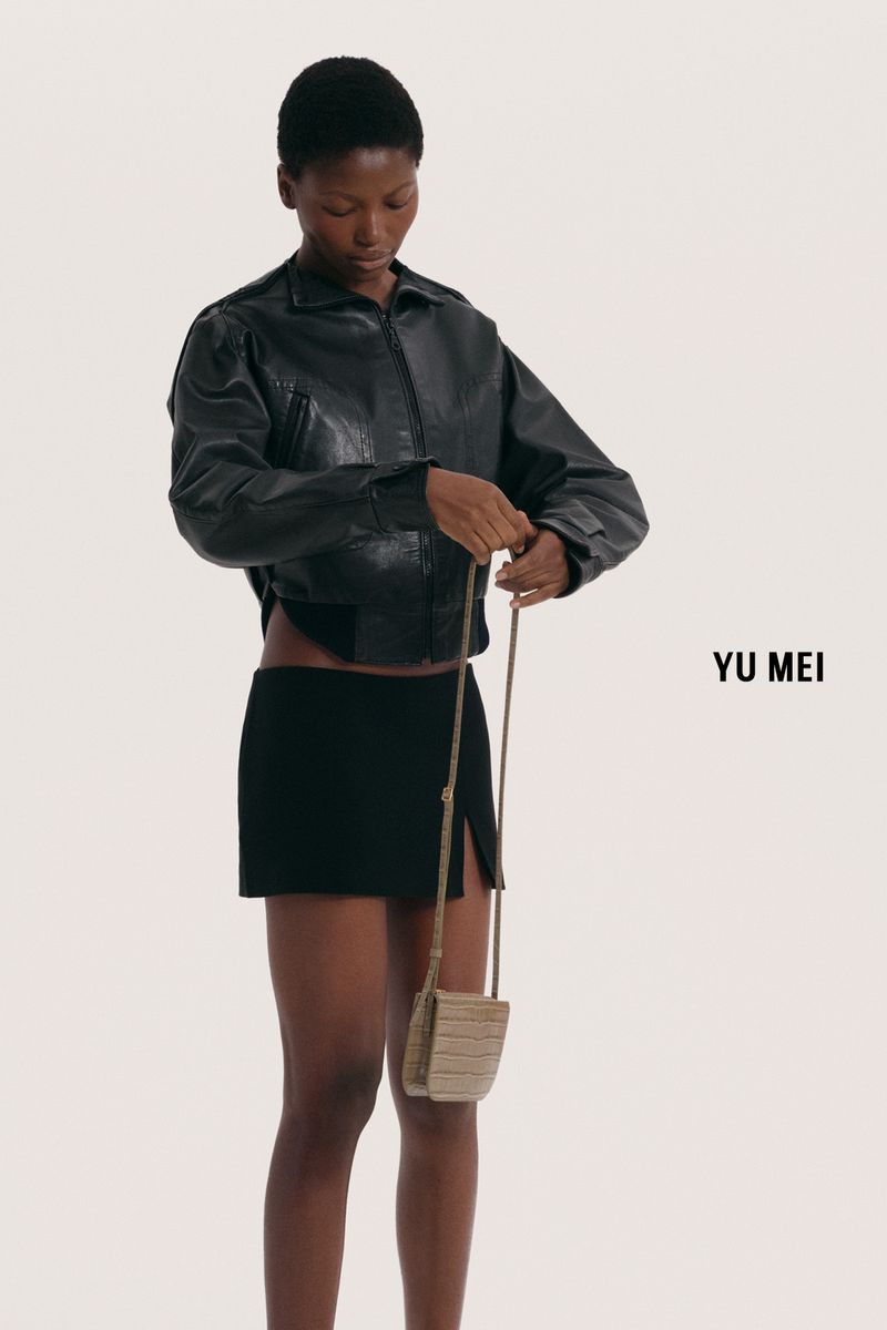 An African-American woman models a black leather jacket, black mini skirt and a tan purse against a white background in a fashion shot.