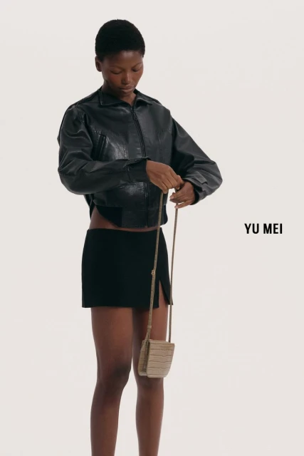 An African-American woman models a black leather jacket, black mini skirt and a tan purse against a white background in a fashion shot.