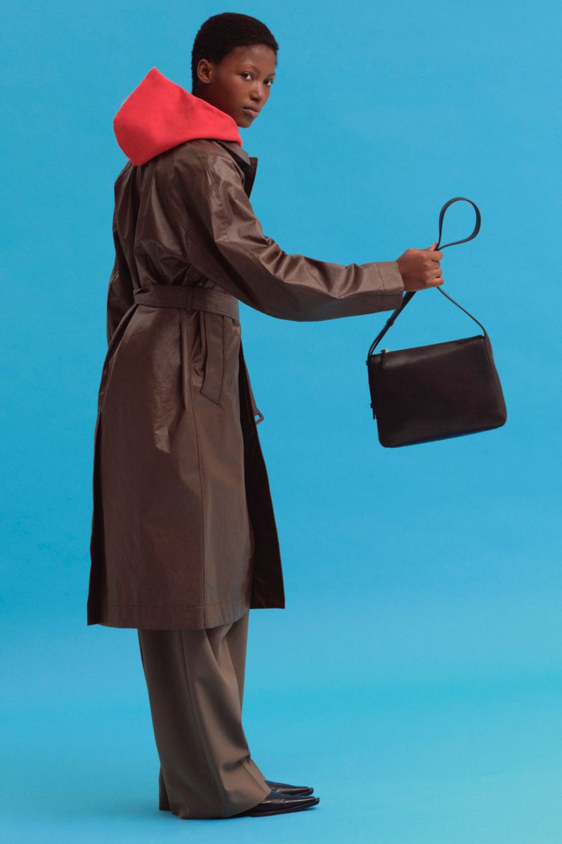 Woman in brown trench coat holds black handbag against blue background. The outfit includes a coral hoodie and dark pants and shoes.