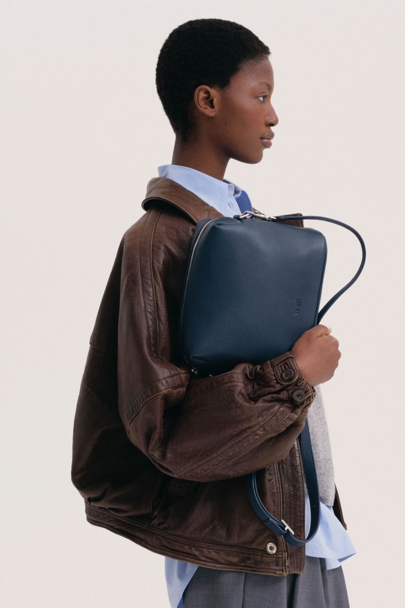 Fashionable dark-skinned woman with a short afro hairstyle wearing a brown bomber jacket holding a dark blue square bag.