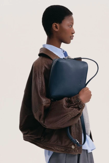 Fashionable dark-skinned woman with a short afro hairstyle wearing a brown bomber jacket holding a dark blue square bag.