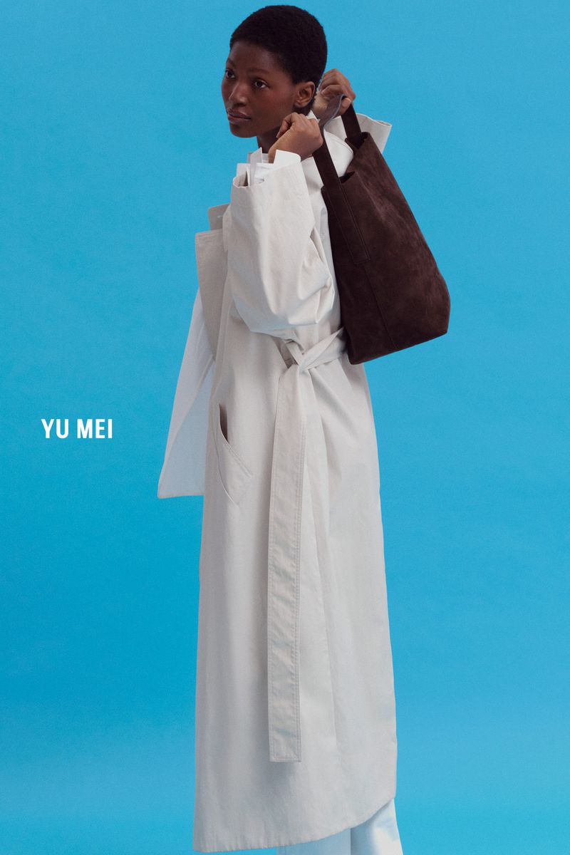 Dark-skinned woman in a light-colored coat holding a brown purse against a light blue background for a fashion advertisement.