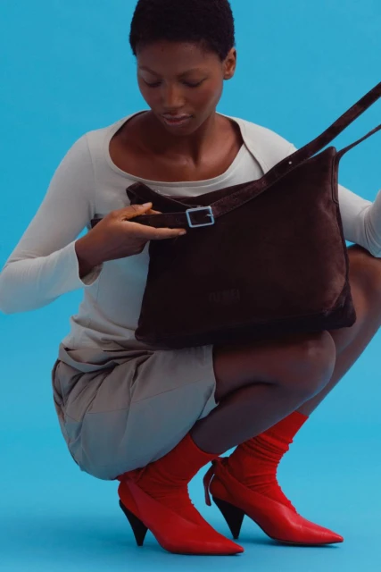 An African-American woman squats in a studio with a brown bag and red heels against a light blue background in a fashion shot.