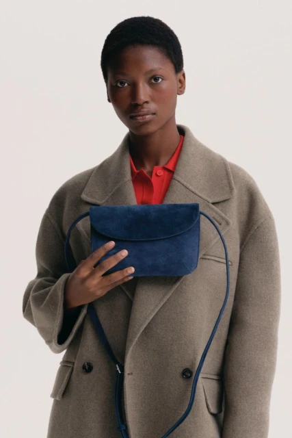 Dark-skinned woman in wool coat holds blue suede bag in studio. Close-up captures stylish fashion accessory and neutral backdrop.