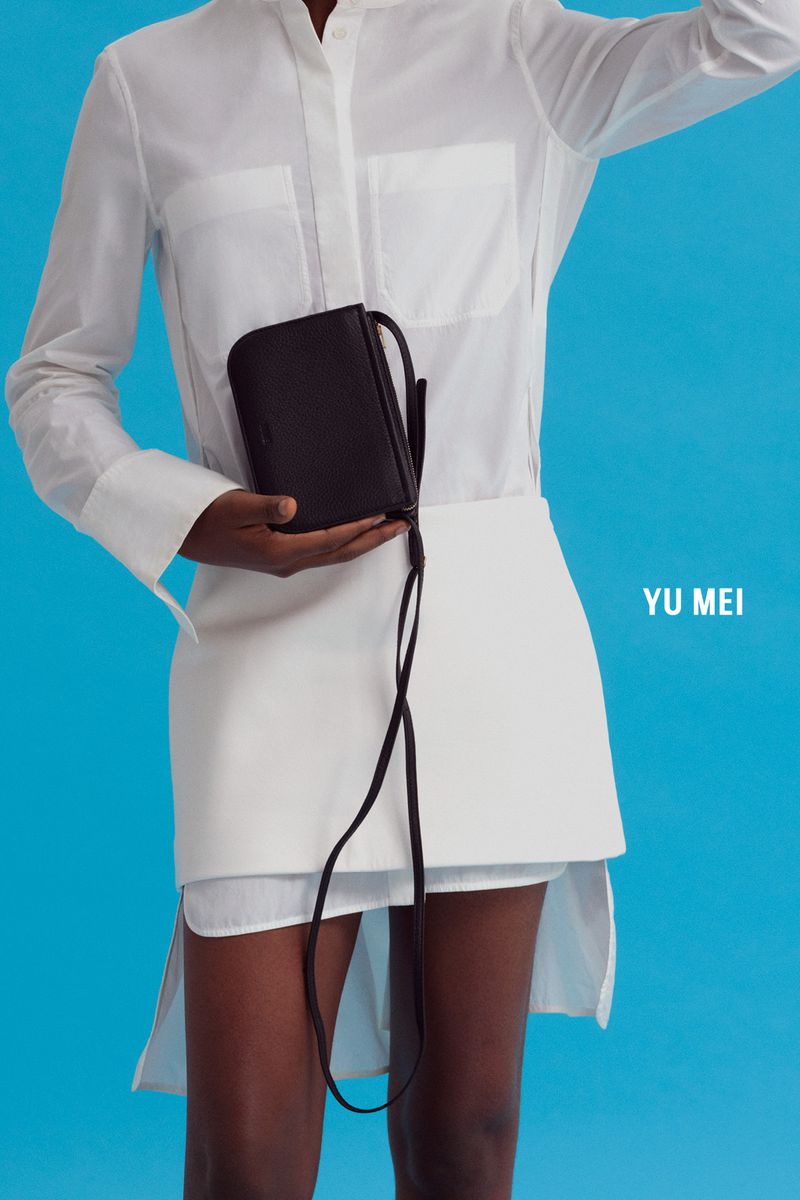 Model wearing a white shirt and skirt holding a dark leather crossbody bag against a bright blue background