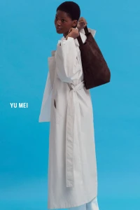 Model wearing a white coat and holding a brown shoulder bag against a bright blue studio background