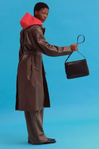 Model in brown trench coat and red hoodie holding a black handbag against a bright blue background
