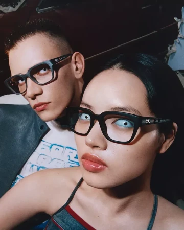 Two people model black-rimmed eyeglasses indoors. The model in the foreground wears blue contact lenses and glossy lipstick. Fashion shot.
