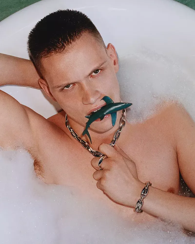 An adult male in a bathtub filled with bubbles, holding a toy shark in his mouth, wearing jewelry, and gazing at the camera.