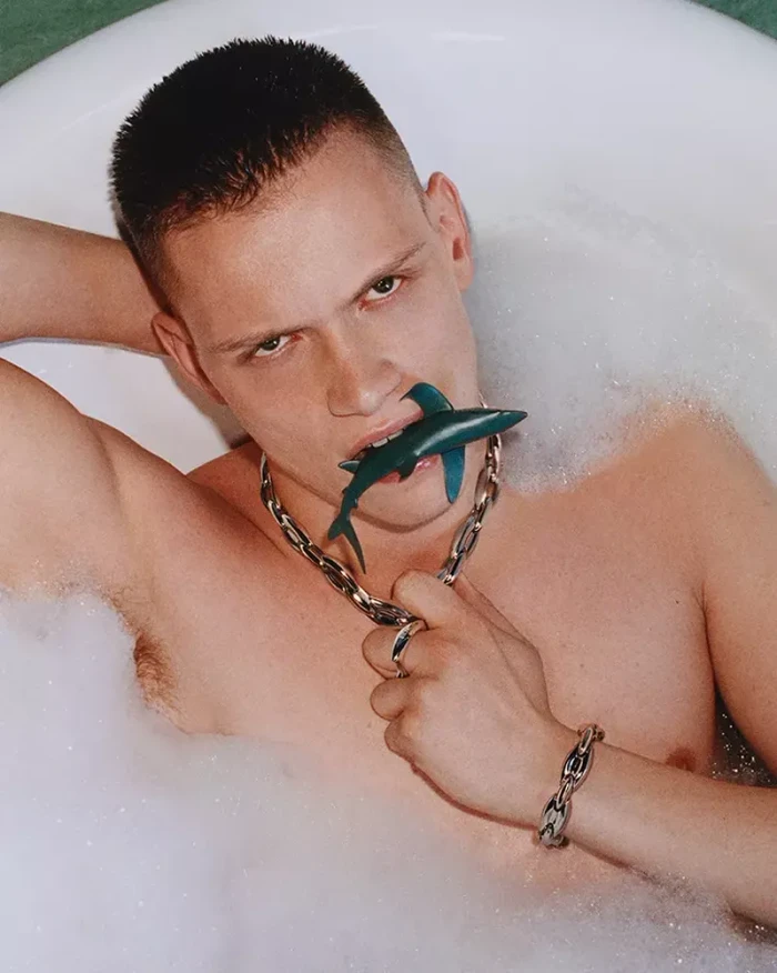 Man with short dark hair in a foam-filled bathtub biting a small toy shark wearing a silver chain