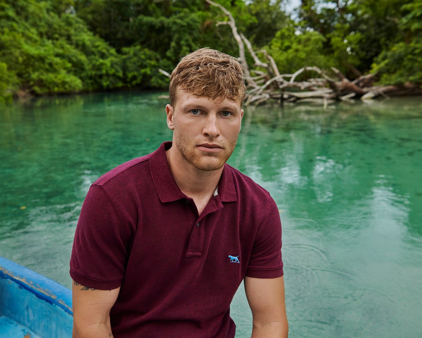 A young adult male sits on a blue boat, looking at the camera with a neutral expression, with green water and trees in the background.