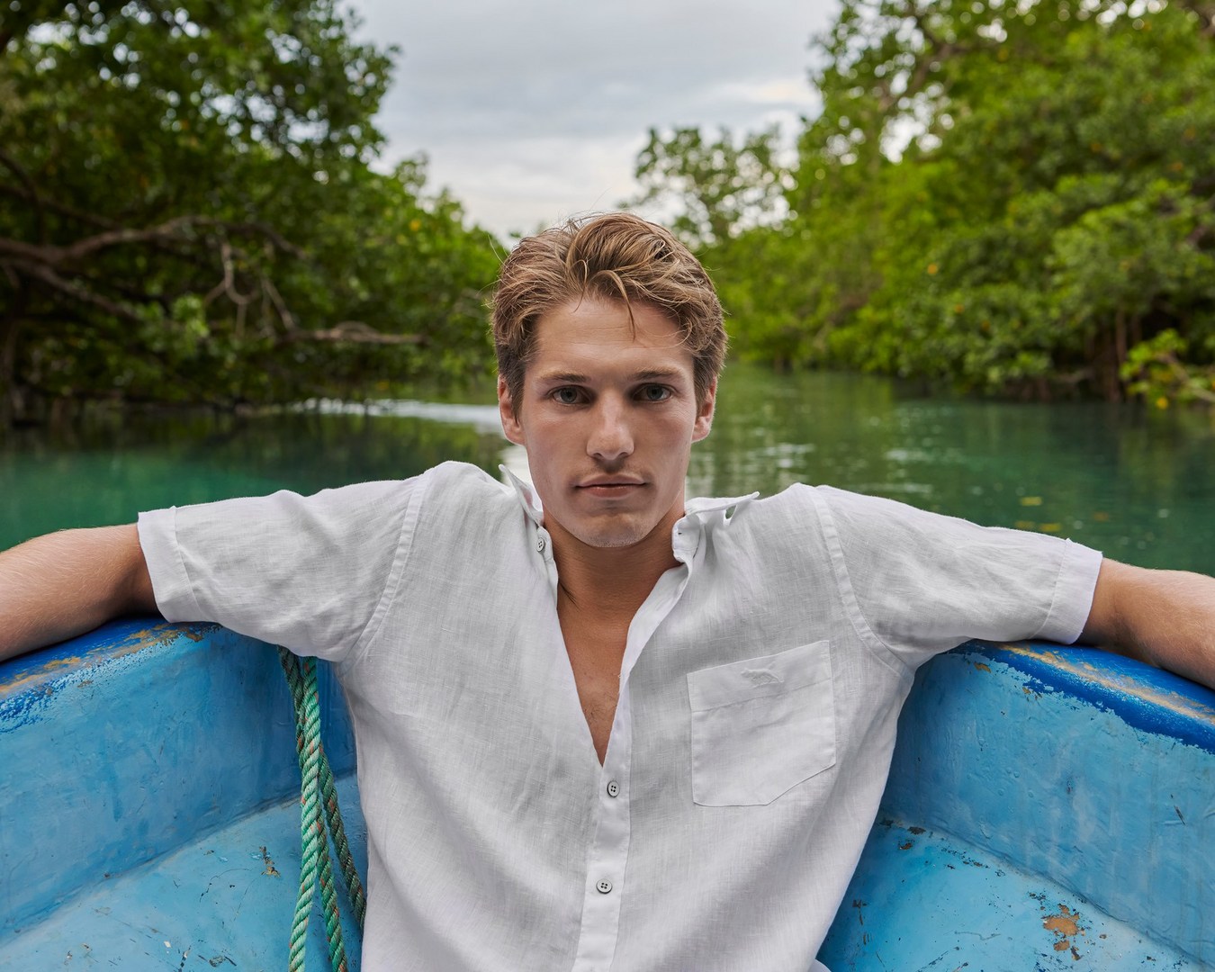 A fair-skinned man with blond hair, wearing a white linen shirt, sits in a blue boat on a river with lush green trees in the background.