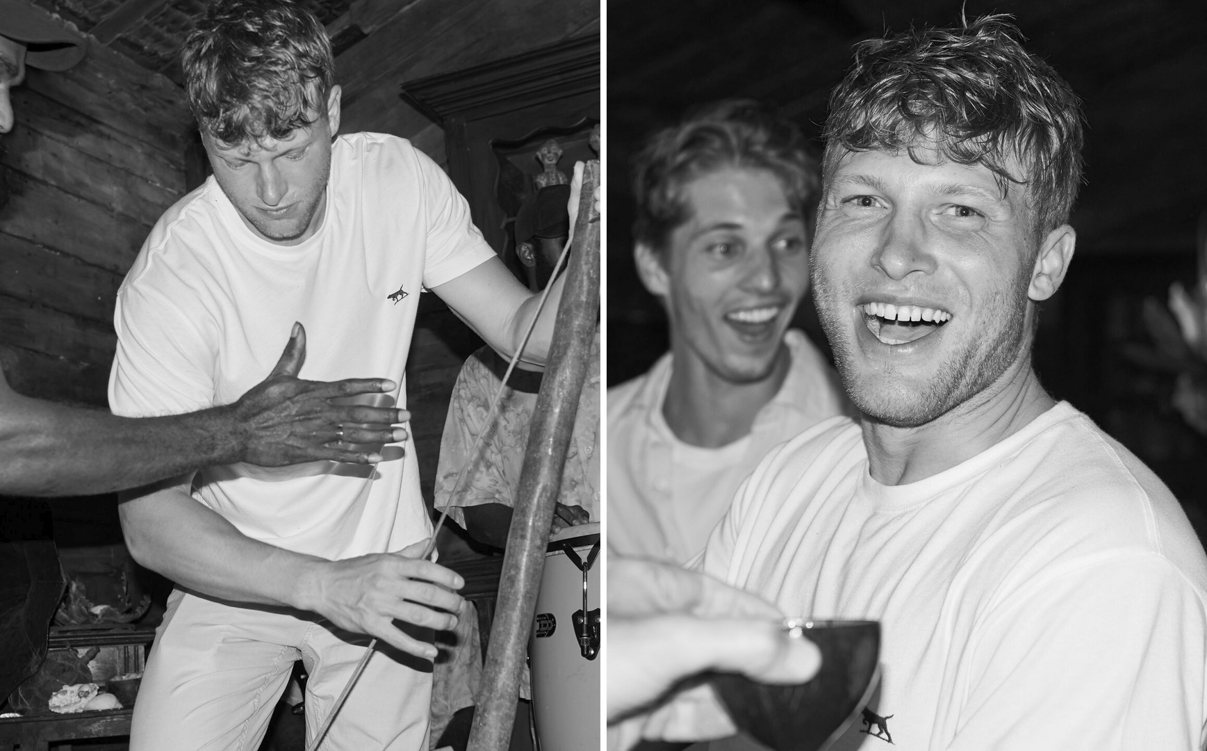 Black and white image of a man playing a musical instrument while another man smiles in the background, indoor setting.